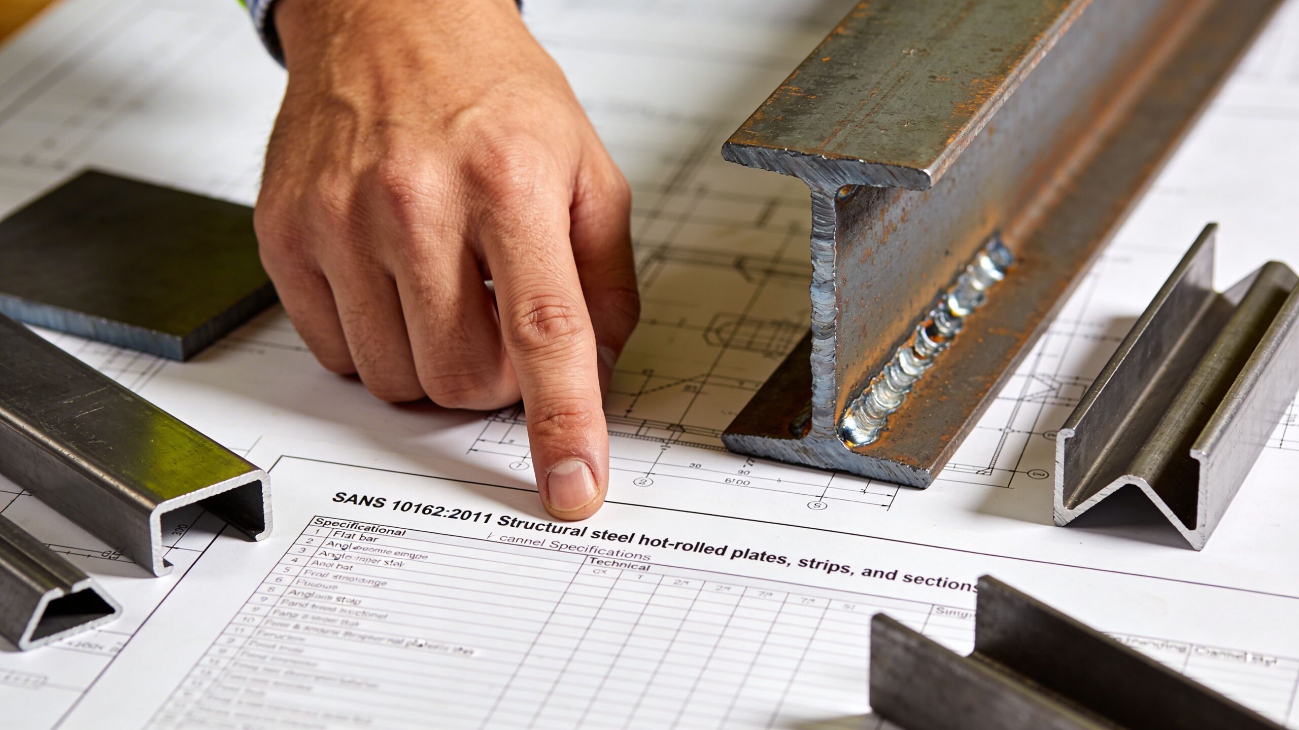 A construction engineer pointing at SANS 10162 steel specification blueprints surrounded by various hot-rolled structural steel sections.